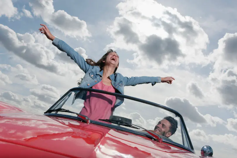 Pareja dentro de coche descapotable, chica con los brazos extendidos y contentos